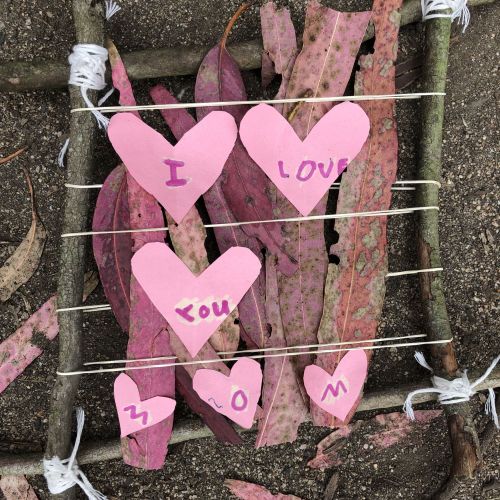 Four sticks are bound together by twine, creating a frame around pink-colored leaves and pink paper hearts that say “I LOVE YOU MOM” in a child’s handwriting.