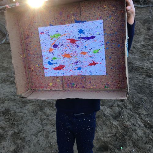 A child holds up a cardboard box that was cut open to reveal a white piece of paper in the middle. The child had splattered the paper with multicolored paint to create a piece of artwork.