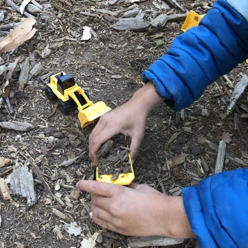 A young child crouches close to the ground, with the camera’s focus on their hands as they fill the scoop of a yellow toy tractor with dirt. Leaves and pieces of bark are scattered around.