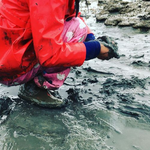 A child in a red jacket, pink heart-patterned pants, and rain boots crouches down to scoop up mud with bare hands.