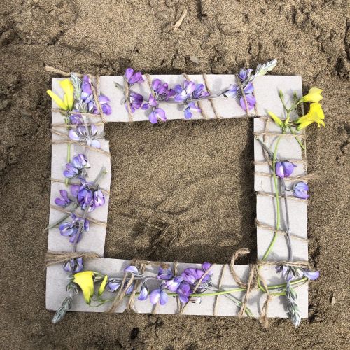 Four pieces of cardboard are held together with twine to make a square white frame, which is decorated with purple and yellow wildflowers tied together.