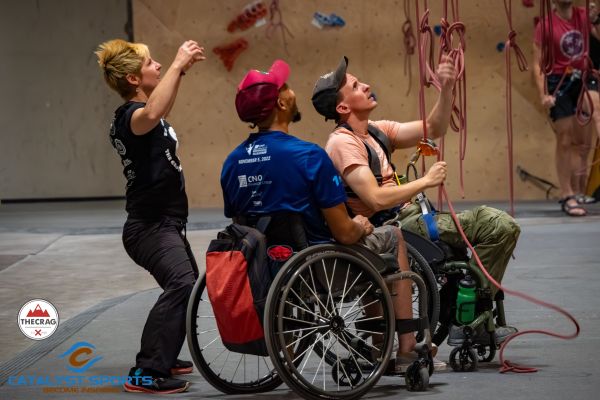 A climber who uses a wheelchair belays another climber during an adaptive climbing program in Murfreesboro, TN. Another person in a wheelchair is watching the belayer and climber on the climbing wall. A volunteer is standing behind the belayer with her arms raised in the air. Ropes hang from the climbing wall in front of the belayer in his wheelchair.