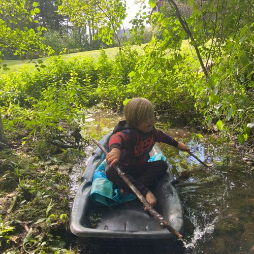 A young boy with blonde hair sits in a small plastic sled in a shallow stream surrounded by lush foliage. Holding a stick and wearing a red shirt and black pants he is enjoying an outdoor adventure.