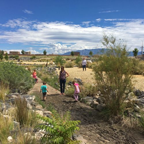 Children explore a muddy path made by a low-flow channel with their feet. One adult is watching the children play and another is walking through the adjacent field.