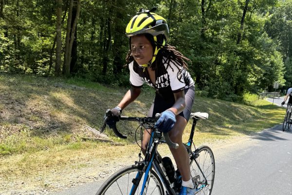 A cyclist rides a bike along a forest trail, which is bordered by thick, green trees that give way to deeper forestry. The cyclist looks ahead to scout the trail before them; they also wear a bright yellow helmet and gloves for safety.