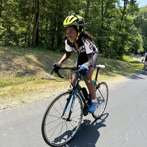 A cyclist rides a bike along a forest trail, which is bordered by thick, green trees that give way to deeper forestry. The cyclist looks ahead to scout the trail before them; they also wear a bright yellow helmet and gloves for safety.