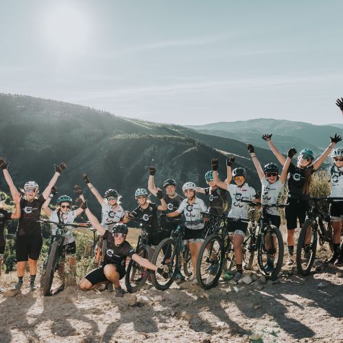 A large group of participants line up for a photo at the edge of a hill. They are all dressed in biking gear and have their arms thrown up in the air as a sign of triumph; some also have bikes by their sides. The background is a beautiful shot of a vast canyon, with rolling green hills that disappear far into the horizon. The clear sky overhead is lit up by a bright sun.