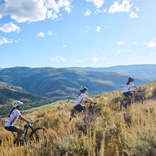 Three participants bike in a line towards the top of a hill. More hills covered by green and brown grass stretch far into the distance, going as far as fading as silhouettes on the horizon. The sun illuminates the sky to reveal a bright blue gradient and clouds strewn across.